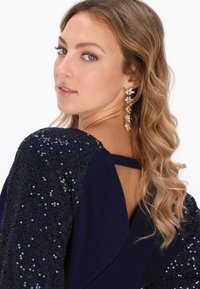 Marineblaues Oberteil mit Paillettenakzenten an den Schultern, das ein Design mit Rückenriemen aufweist. Lockige Haare und hängende Ohrringe ergänzen den Look.