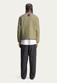 Oliwkowa pikowana kurtka o fakturowanym wzorze, z czarnym wykończeniem i widocznym logo przy dekolcie, zestawiona z czarnymi spodniami o luźnym kroju.