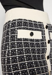 Černobílá kostkovaná sukně s proužkovaným krémovým pasem, obsahující boční kapsu a černé knoflíky na přední části. Hladká textura látky.