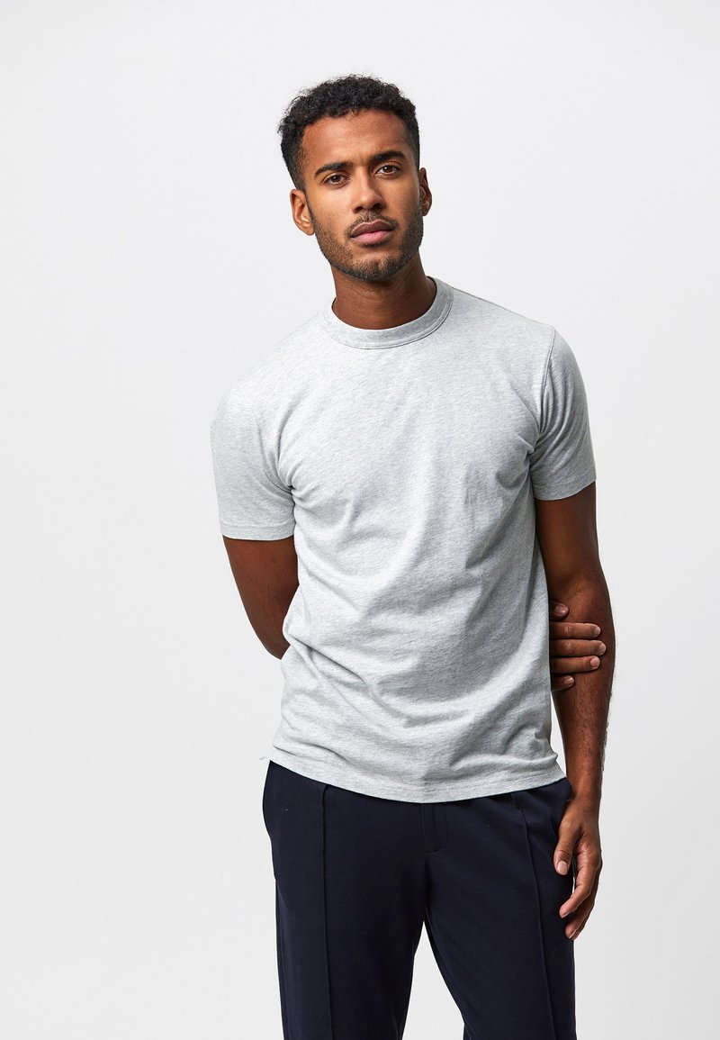 Mężczyzna o krótkich kręconych włosach, ubrany w jasnoszarą koszulkę i ciemne spodnie, stoi na tle jednolitego jasnego tła.