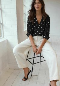 Schwarz gepunktete Bluse mit V-Ausschnitt, kombiniert mit weiten weißen Hosen. Das Outfit wird durch schwarze Absatzsandalen ergänzt.