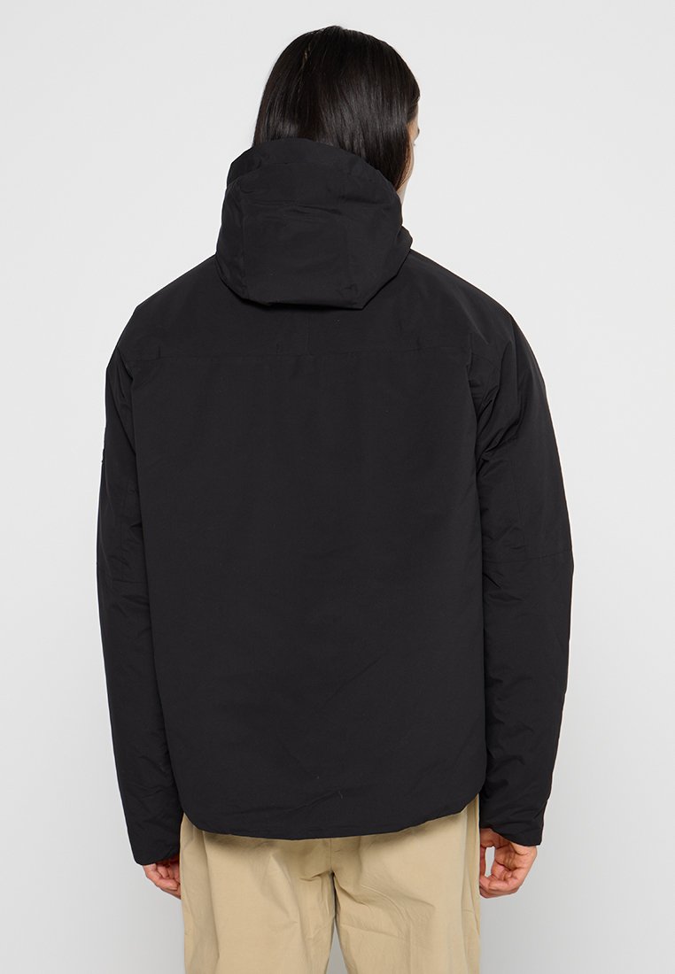 Giacca nera imbottita con cappuccio staccabile e taglio dritto. Texture liscia e senza motivi visibili. Vista posteriore, indossata con pantaloni beige.