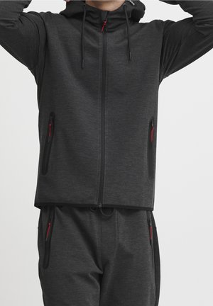 Mann trägt dunkelgrauen Kapuzenpullover mit Reißverschluss und passende Jogginghose mit roten Reißverschlussanhängern, zieht die Kapuze hoch, Ansicht von Hals bis Oberschenkel.