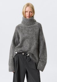Pulover oversized de culoare gri deschis, cu guler mare, fabricat dintr-un material moale și pufos. Are mâneci largi și o croială relaxată. Este asortat cu pantaloni gri închis și o curea.