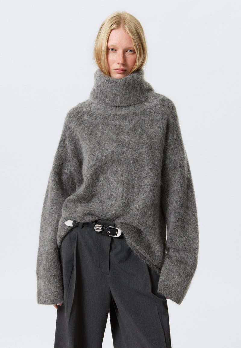Pulover oversized de culoare gri deschis, cu guler mare, fabricat dintr-un material moale și pufos. Are mâneci largi și o croială relaxată. Este asortat cu pantaloni gri închis și o curea.