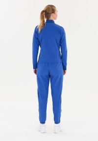 Kvinde stående med ryggen til iført en matchende blå træningsdragt og hvide sneakers på en helt hvid baggrund.