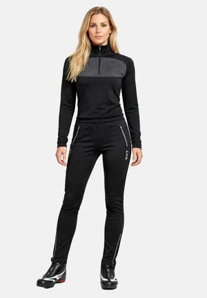 Frau steht vor weißem Hintergrund in schwarzer Sportjacke mit Reißverschluss, dazu passende Hose mit Reißverschlusstaschen und schwarzen Sportschuhen.