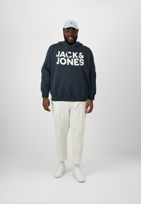 Navy hoodie met grote witte opdruk "JACK & JONES", kangoeroezak, witte tapering broek en lichtblauwe pet, gecombineerd met witte sneakers.