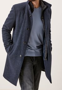 Cappotto navy in tessuto testurizzato, con chiusura a bottoni e foderato in nero. Indossato sopra un maglione azzurro chiaro, abbinato a jeans scuri.