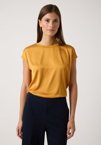 Senfgelb kurzärmlige Bluse mit rundem Halsausschnitt, glatter Textur und lockerer Passform, kombiniert mit dunkelblauen Hosen.