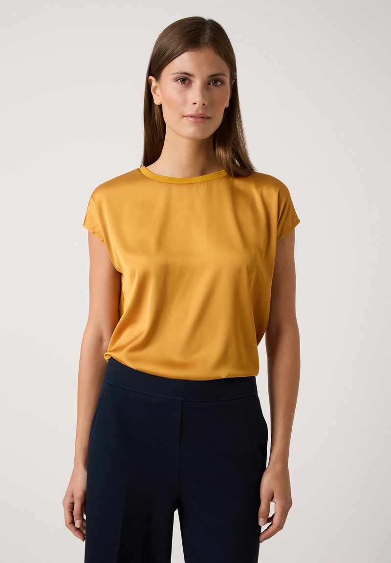 Senfgelb kurzärmlige Bluse mit rundem Halsausschnitt, glatter Textur und lockerer Passform, kombiniert mit dunkelblauen Hosen.