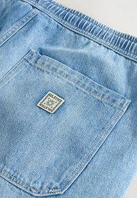 Pânză de denim de culoare albastru deschis, cu un manșon elastic, având o bătaie la spate și un etichete cusută inscripționată "Solstice". Textură netedă cu cusături.
