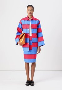 Gestreiftes Blazer- und Rock-Set aus rotem und blauem Stoff, mit weiten Ärmeln, Knopfverschlüssen und einer strukturierten Oberfläche. Hält eine orangefarbene Handtasche.