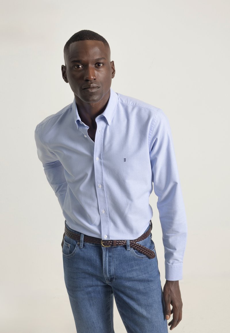Camisa de manga larga azul claro con cuello abotonado, combinada con unos pantalones vaqueros azul oscuro y un cinturón trenzado marrón, destacando un ajuste clásico.