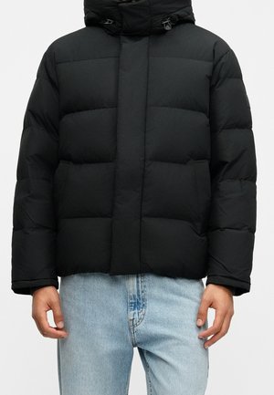 Mężczyzna ubrany w czarną kurtkę puchową z kapturem i jasnoniebieskie dżinsy, stojący na jednolitym białym tle.