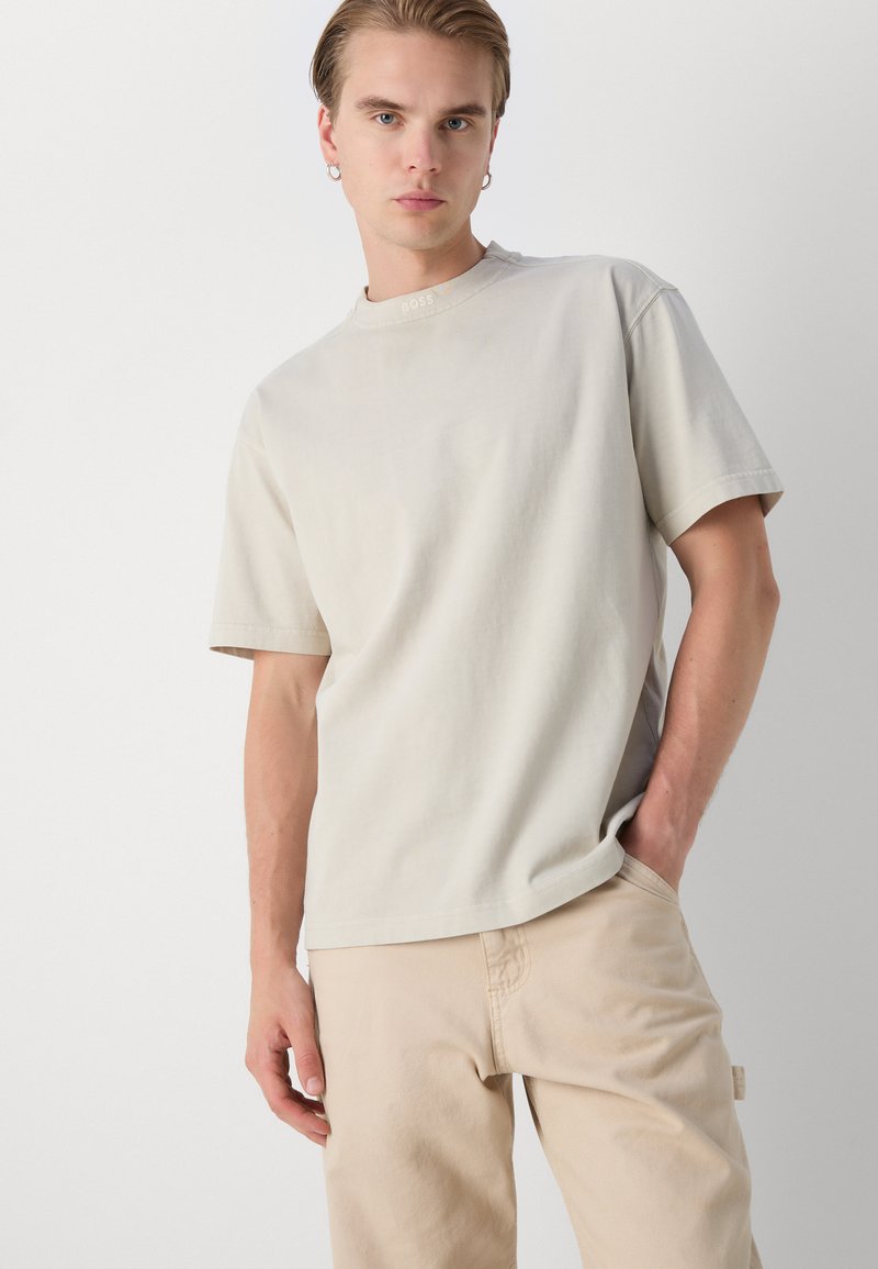 Lichtbeige katoenen T-shirt met een ronde halslijn en korte mouwen, met een klein reliëf merkembleem langs de kraag.