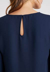 Námořnická modrá halenka s klíčovou miskou na zádech, vyrobená z měkké a hladké látky s čistým výstřihem a uvolněnými rukávy.