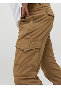 Personne portant un pantalon cargo marron avec poches à rabat, la main insérée dans la poche avant, sur un fond clair uni.