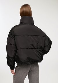 Giacca a vento nera con colletto alto, design oversize e polsini a coste. Tessuto liscio e lucido con cuciture orizzontali per l'isolamento.