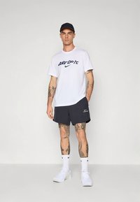 Férfi Nike ruházat: fehér póló kék "Just Do It" felirattal, tengerészkék rövidnadrág, fehér zokni és fehér sportcipő. Jól látható tetoválások a karokon.