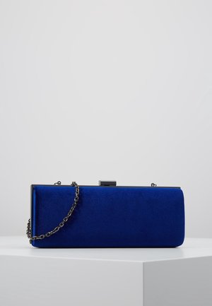 Pochette rectangulaire en velours bleu royal avec cadre métallique noir et bandoulière chaîne, présentée sur un piédestal blanc sur un fond uni.