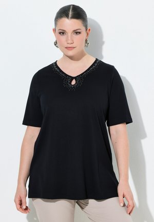 Donna con capelli scuri raccolti indossa una maglietta nera a maniche corte con scollo a goccia decorato e pantaloni beige, in piedi contro un muro bianco.