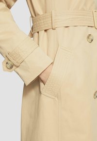 Beige trenchcoat met een riem in de taille, met knoopmanchetten en zijzakken. Heeft een gestructureerde stof en stikselsdetails op de mouwen.