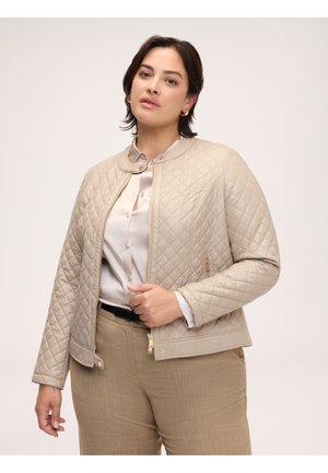 Kvinde iført en beige quiltet jakke, lys bluse og lysebrune bukser, stående mod en ensfarvet lys baggrund.
