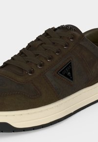 Καφέ sneaker Guess με σκούρα καφέ κορδόνια, μαύρο τρίγωνο λογότυπο στο πλάι και λευκή σόλα με μαύρο πέλμα στο κάτω μέρος.