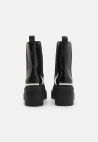 Botines de charol negro con tacón grueso, paneles laterales elásticos y detalle de marca en el tacón. Textura suave y diseño elegante.