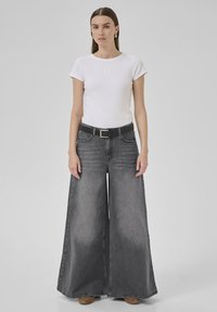 Woman wearing white fitted short-sleeve top, wide-leg high-waisted gray jeans with black belt, and tan shoes, standing against plain background.