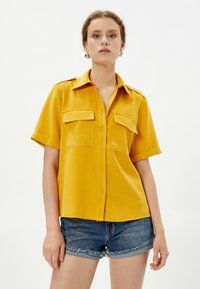 Camicia gialla senape a maniche corte con bottoni frontali, due tasche sul petto e spalline sulle spalle, realizzata in un tessuto morbido. Indossata con pantaloni corti in denim.