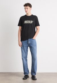 T-shirt en coton noir avec le logo "BOSS" en blanc, associé à un jean en denim bleu clair et des chaussures noires. Tenue simple et décontractée avec une coupe standard.