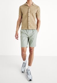 Camicia beige a maniche corte in maglia con motivo a onde, abbinata a pantaloni corti verde chiaro. Sneakers grigie con dettagli bianchi e calzini bianchi.