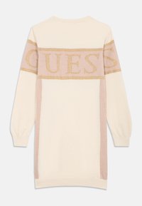 Pletené šaty v krémové a světle růžové barvě, s nápisem "GUESS" v zlatém třpytu na přední straně, s dlouhými rukávy a žebrovanými manžetami.