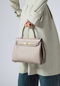 Harmaa nahkahandbag, jolla on jämäkkä muoto, kultaiset yksityiskohdat ja sileä, teksturoitu pinta. Kantaa henkilö, jolla on päällään vaalea takki.