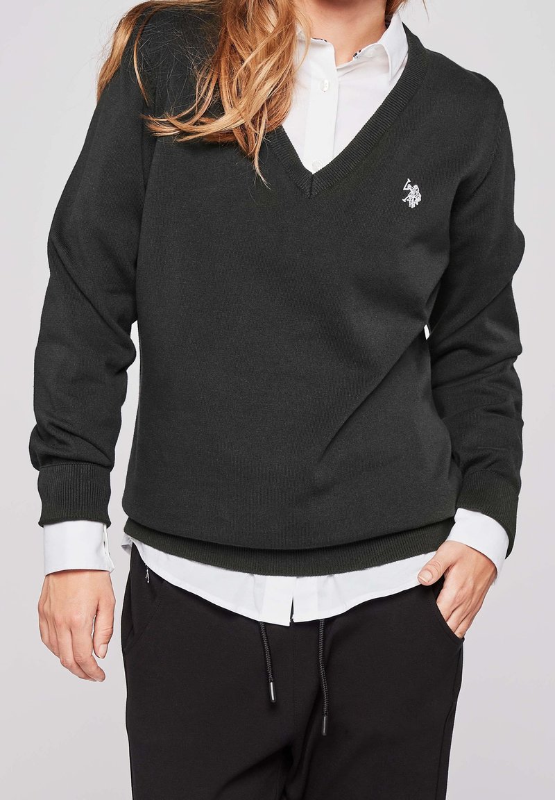Femme portant un pull noir à encolure en V par-dessus une chemise blanche boutonnée avec un pantalon noir, mains détendues, cadrée au niveau du cou et des hanches.