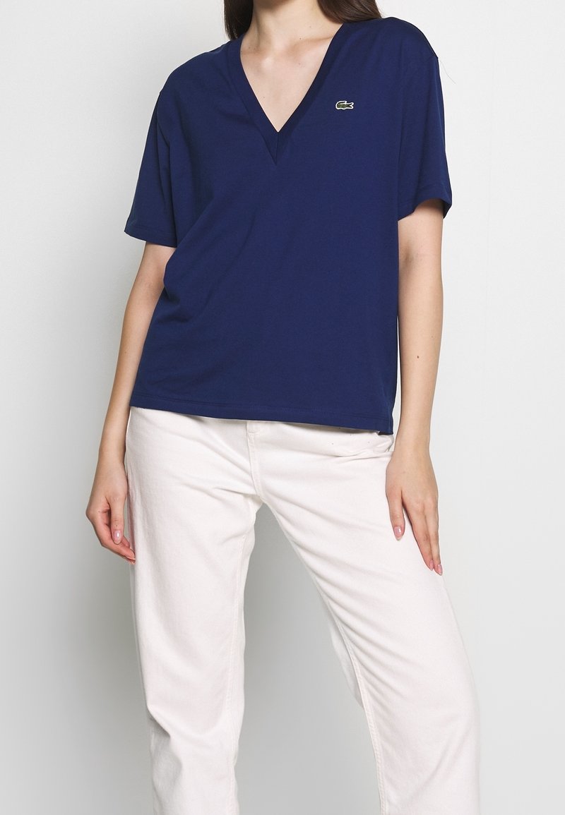 Navy V-neck t-shirt made of cotton, featuring short sleeves and a small logo accent on the upper left. Paired with white straight-leg pants.