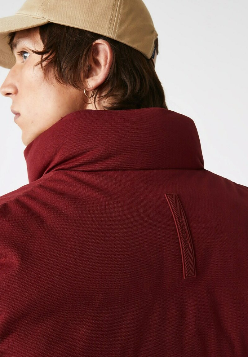 Bordeauxfarbene Jacke mit hohem Kragen, strukturiertem Stoff und kontrastierendem Logo-Detail auf dem Rücken, kombiniert mit einer hellbeigen Kappe.