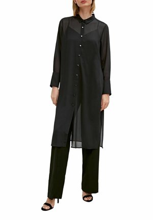 Vrouw draagt een lange, doorschijnende zwarte blouse met knopen over zwarte wijde broek en zwarte open teen hakken, staand tegen een witte achtergrond.