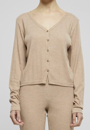 Beige gerippte Strickjacke mit Knöpfen, getragen über passenden gerippten Strickhosen, dargestellt von Hals bis Hüfte vor neutralem Hintergrund.