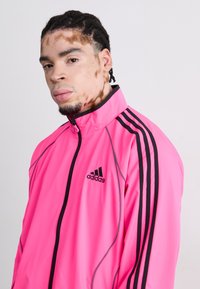 Pink Adidas track jacket with black zipper and three black stripes along sleeves. Features a high collar and lightweight, smooth texture.