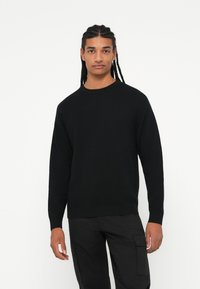 Maglione nero lavorato a maglia con scollo rotondo, maniche lunghe, tessuto testurizzato e polsini a costine. Indossato con pantaloni cargo neri coordinati.