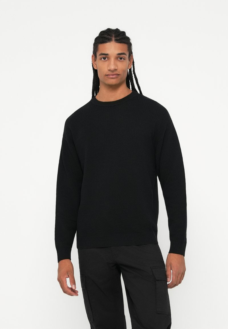 Maglione nero lavorato a maglia con scollo rotondo, maniche lunghe, tessuto testurizzato e polsini a costine. Indossato con pantaloni cargo neri coordinati.