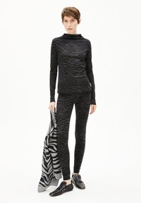Zwarte longsleeve-top en bijpassende legging met een zebra-streep patroon; model houdt een gestructureerde grijs en zwart gestreepte trui vast. Zwarte schoenen met studs.