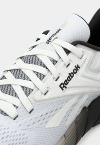 Pantofii sport albi cu partea superioară din plasă, accente gri și guler căptușit. Prezintă un logo negru, șireturi plate și o talpă texturată cu segmente multicolore.