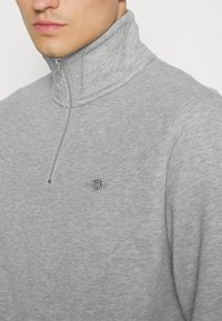 Gros plan d'une personne portant un pull gris clair à fermeture éclair quart de longueur avec un petit logo écusson brodé sur la poitrine.