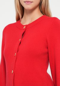 Red knitted cardigan with a round neckline, gold buttons, and side pockets. Soft texture and fitted design for a streamlined look.