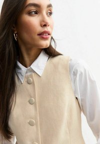 Chaleco sin mangas beige con cinco botones redondos, tejido texturizado, combinado con una camisa blanca de cuello. Cabello cayendo sobre los hombros.