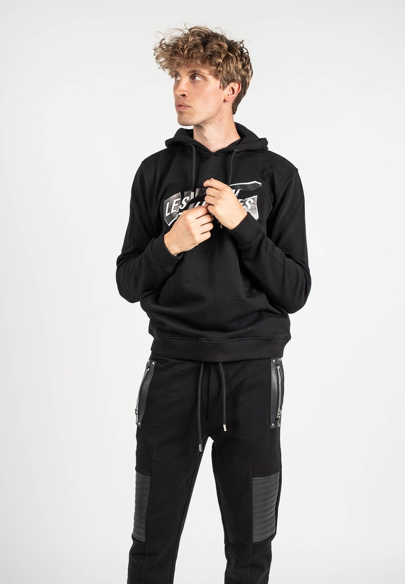 Sudadera negra con un logo en el centro, cordones ajustables y puños acanalados. Combinada con pantalones jogger negros que cuentan con bolsillos con cremallera y paneles texturizados.